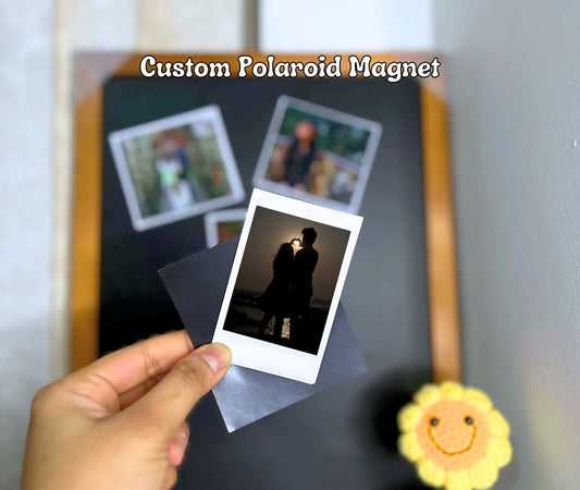 A hand holding a Polaroid-style custom photo magnet featuring a silhouette of a couple at sunset, with a black magnetic board in the background.